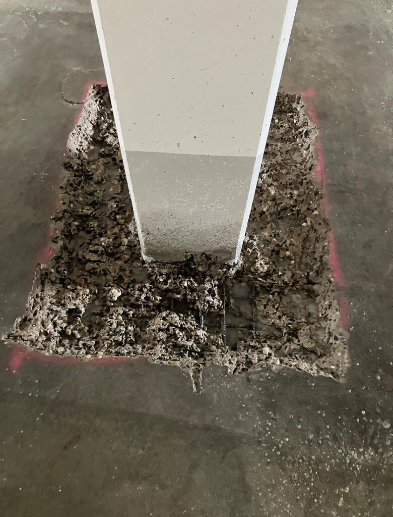 Im Mittelpunkt steht ein rechteckiger Betonpfeiler, der senkrecht aus einer quadratischen Aussparung im Boden ragt. Die Aussparung ist mit grobem, grauem Erdmaterial gefüllt und von einer roten Markierung umrandet. Der Boden besteht aus glattem, dunklem Beton.