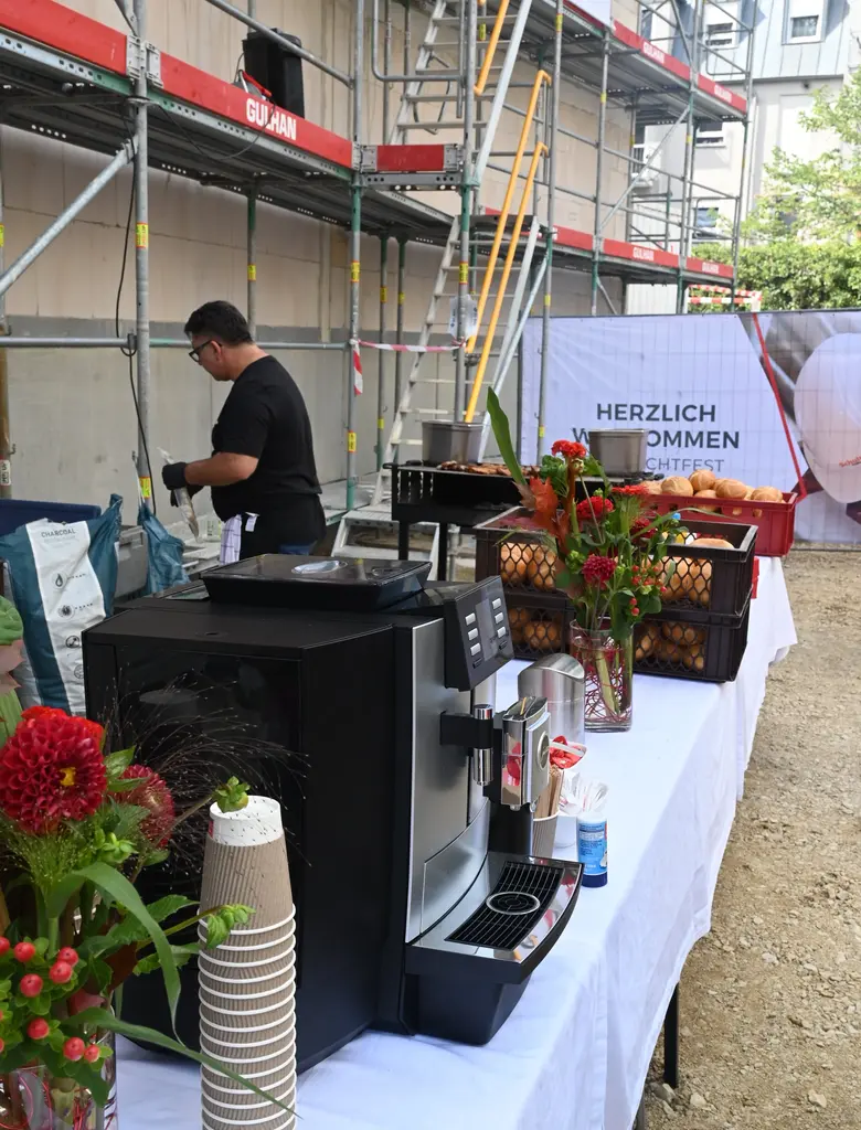 Im Vordergrund steht eine silberne Kaffeemaschine auf einem Tisch, daneben gestapelte Pappbecher mit braun-weißem Muster. Der Tisch ist mit roten Blumen in Glasvasen und mehreren Körben mit Brötchen dekoriert. Im Hintergrund befindet sich ein Mann in dunkler Kleidung, der an einem weiteren Tisch arbeitet. Dahinter sind Baugerüste und eine Wand zu sehen. Ein großes Banner mit der Aufschrift 'HERZLICH WILLKOMMEN' steht rechts neben dem Tisch. Die Szene spielt im Freien.