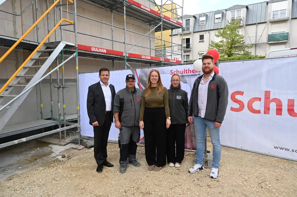 Fünf Personen stehen nebeneinander auf einem Schotterboden vor einer Baustelle. Im Hintergrund sind ein Baugerüst, ein teilweise verputztes Gebäude und ein großes Banner mit roter Schrift zu sehen. Die Personen tragen unterschiedliche Kleidung: Anzüge, Arbeitskleidung und Freizeitoutfits. Alle blicken in Richtung Kamera und lächeln.