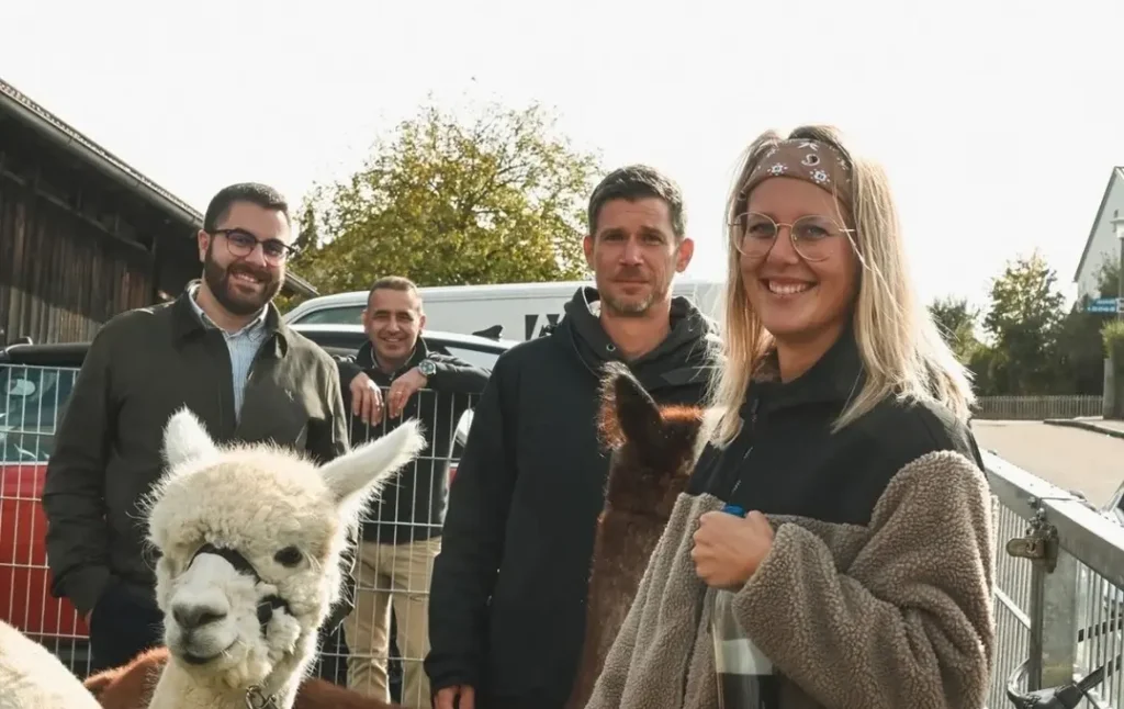 Im Vordergrund steht eine junge Frau mit langen blonden Haaren, Brille und Stirnband, die lächelnd in die Kamera blickt. Sie trägt eine zweifarbige Fleecejacke und hält eine Flasche in der Hand. Neben ihr befindet sich ein Alpaka mit hellem, lockigem Fell und dunklen Flecken im Gesicht. Im Hintergrund stehen drei Männer, die lächeln oder neutral schauen. Sie tragen Freizeitkleidung und stehen vor einem Metallzaun. Dahinter sind ein Transporter, ein rotes Auto und ein Gebäude mit Holzfassade zu sehen. Im Hintergrund wachsen Bäume, der Himmel ist hell.