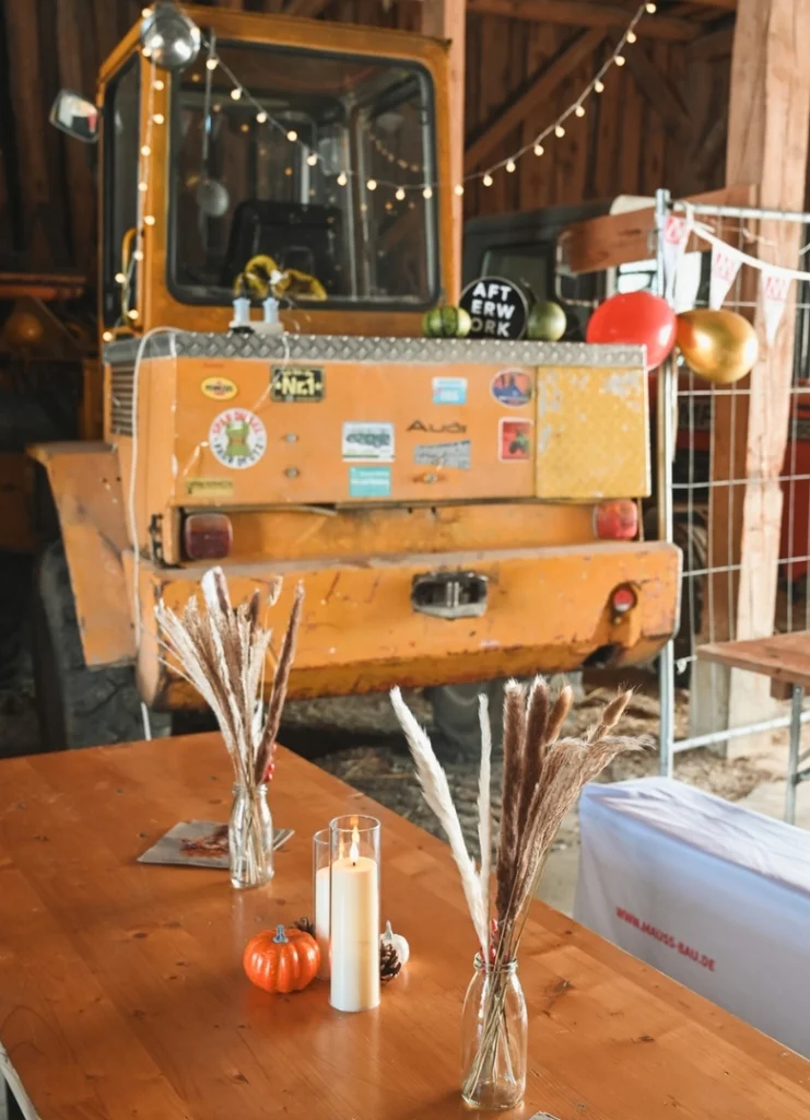 Im Vordergrund steht ein Holztisch, auf dem mehrere Glasvasen mit getrockneten Gräsern, eine Kerze im Glas und eine kleine orangefarbene Kürbisdekoration arrangiert sind. Im Hintergrund befindet sich ein gelber Bagger mit verschiedenen Aufklebern auf der Rückseite. Darüber hängen Lichterketten und einige goldene und rote Luftballons. Die Szene spielt in einer Scheune mit Holzverkleidung.
