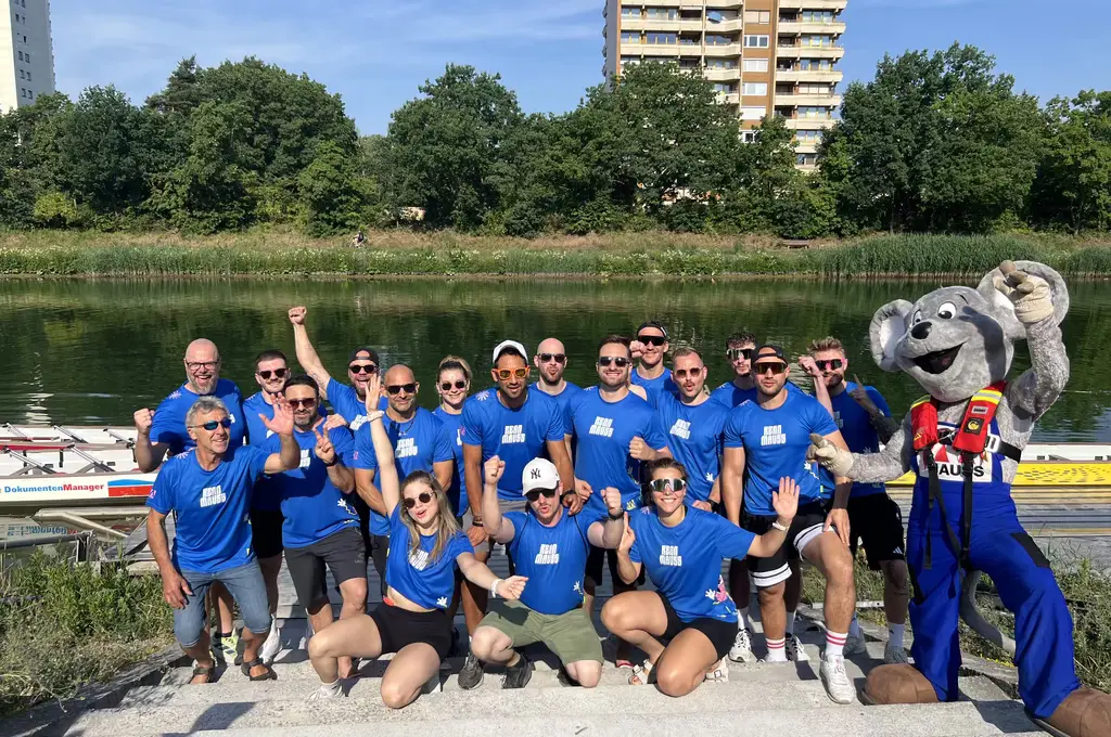Eine Gruppe von Menschen steht am Ufer eines Gewässers und trägt einheitliche, blaue T-Shirts. Sie posieren fröhlich mit erhobenen Armen und lächeln in die Kamera. Rechts steht eine Person im großen, grauen Maskottchenkostüm mit Tierkopf, blauer Hose und roter Weste. Im Hintergrund sind Bäume, ein Schilfgürtel und mehrstöckige Wohngebäude zu sehen. Im Wasser liegen mehrere schmale Boote.