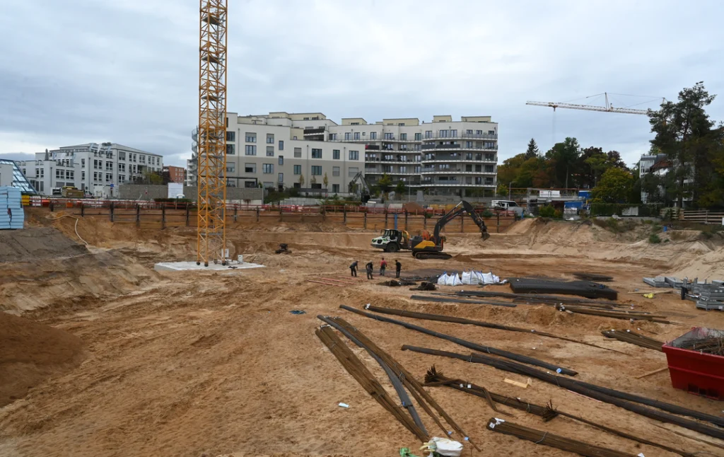 Eine große Baustelle mit sandigem Boden und mehreren Baugeräten. Im Vordergrund liegen lange Rohre und Baumaterialien. Links steht ein gelber Baukran, im Hintergrund sind moderne mehrstöckige Wohngebäude zu sehen. Einige Personen arbeiten in der Mitte des Baugeländes, daneben befindet sich ein Bagger. Der Himmel ist bewölkt.
