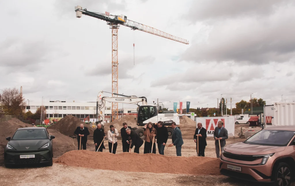 Eine Gruppe von Menschen steht auf einer Baustelle und hält Spaten in den Händen, als würde ein symbolischer erster Spatenstich stattfinden. Im Hintergrund sind ein großer Baukran, ein Bagger, mehrere Container und ein Firmenlogo auf einem Bauzaun zu sehen. Links und rechts im Vordergrund stehen zwei moderne Autos. Der Himmel ist bewölkt.