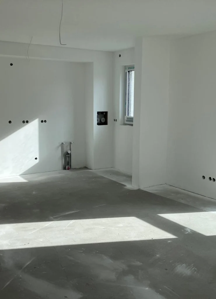 Heller Raum mit weiß gestrichenen Wänden und grauem Betonboden. Mehrere Steckdosen und Wandöffnungen sind sichtbar, ebenso ein kleines Fenster mit silbernem Rahmen. An einer Wand befindet sich eine freiliegende Wasserleitung und eine schwarze Wandplatte. Sonnenlicht fällt durch das Fenster und erzeugt markante Lichtflächen auf dem Boden.