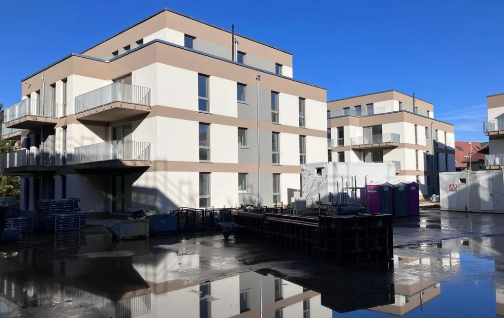 Mehrgeschossige, moderne Wohngebäude mit flachen Dächern und hellen Fassaden stehen auf einem asphaltierten Gelände. Die Gebäude haben große Fenster und mehrere Balkone mit Metallgeländern. Im Vordergrund befinden sich Pfützen, Bauzäune, Baumaterialien und mobile Toiletten, was auf eine Baustellensituation hindeutet. Der Himmel ist klar und blau.