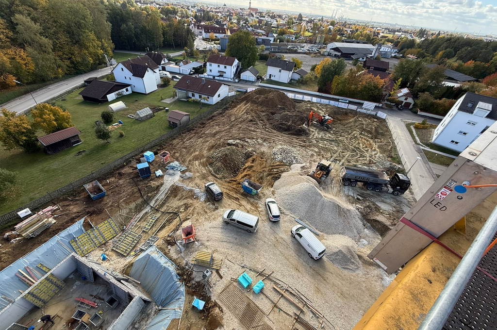 Blick von oben auf eine Baustelle mit mehreren Fahrzeugen, darunter Transporter, ein Bagger und ein Lastwagen. Im Vordergrund sind Baumaterialien, Bauzäune und Baugruben zu sehen. Im Hintergrund befinden sich Wohnhäuser mit Gärten, Bäume mit herbstlicher Färbung und eine weitläufige Siedlung. Die Szene ist bei Tageslicht aufgenommen, der Himmel ist leicht bewölkt.